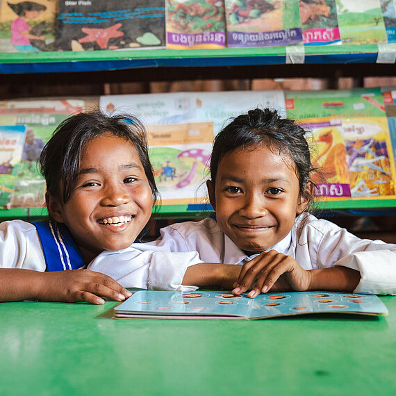 Zwei lächelnde Mädchen in Schuluniform sitzen an einem grünen Tisch mit einem aufgeschlagenen Buch, im Hintergrund ein Regal mit bunten Büchern, aufgenommen in einer Schule in Kambodscha.
