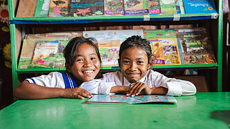 Zwei lächelnde Mädchen in Schuluniform sitzen an einem grünen Tisch mit einem aufgeschlagenen Buch, im Hintergrund ein Regal mit bunten Büchern, aufgenommen in einer Schule in Kambodscha.