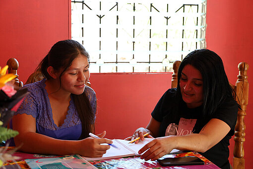 Zwei Frauen sitzen an einem Tisch und sind über Schularbeiten gebeugt