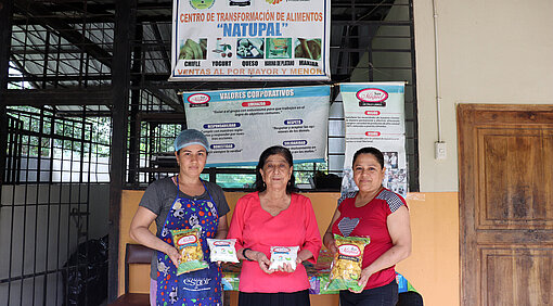 Drei Frauen stehen vor einem großen Schild, auf dem "Centro de transformación de alimentos Natupal" steht
