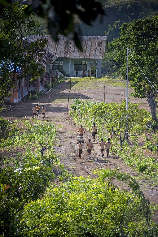 Kinder auf dem Weg zur Grundschule im indonesischen Bezirk Nagekeo Mehrere flache Gebäude stehen an einem Berghang zwischen Bäumen