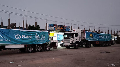 Zwei LKW mit Hilfslieferungen stehen in der Abenddämmerung. Auf ihnen sind große Transparente mit der Aufschrift "Plan International" angebracht.