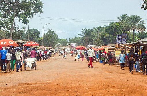 Mensche laufen über eine rostrote Sandpiste, an der kleine Läden aufgebaut sind