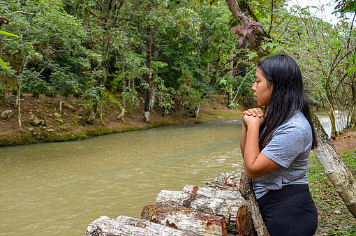 Anahi steht mit verschränkten Händen am Flussufer und blickt auf das Wasser