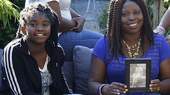 Fatu, rechts neben einer ihrer Töchter, hält ein Foto des Paares hoch, das sie bis zu ihrem 18. Lebensjahr durch eine Patenschaft unterstützt hat.