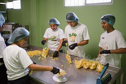 Mehrere Personen sind in einem Produktionsraum mit Haarnetzen und Schutzkleidung zu sehen. Sie verpacken gelbe Chips an einem Edelstahltisch. Die Szene zeigt ein Arbeitsteam bei der Lebensmittelverarbeitung.