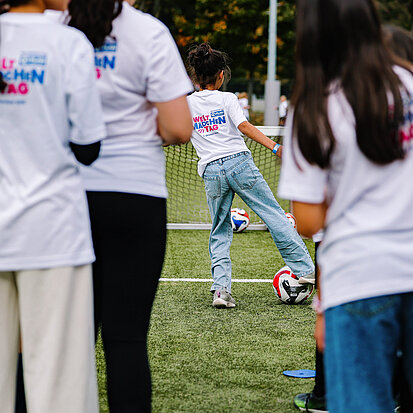 Eine Gruppe Mädchen beobachten ein Mädchen, die einen Fußball auf ein kleines Tor schießt. Sie tragen T-Shirts mit der Aufschrift "Weltmädchentag".