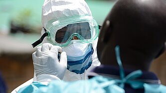 Zwei Personen, davon eine im Vordergrund unscharf, die andere im Hintergrund trägt Schutzkleidung, einschließlich Schutzbrille, Maske und Haube.