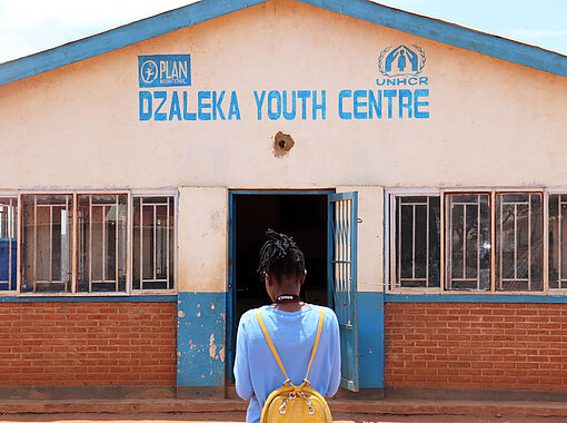 Clementine steht mit dem Rücken zur Kamera. Vor ihr ist ein Gebäude, auf dem Dzaleka Youth Centre steht. 