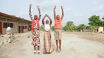 Drei Mädchen stehen auf einem sandigen Vorplatz einer Schule und springen in die Luft