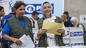 Zwei Mitarbeiterinnen von Plan International stehen in einem Lagerraum mit Hilfspaketen und Papieren in der Hand. Im Hintergrund sind weitere Helfer:innen und Kartons mit dem Plan-Logo zu sehen.