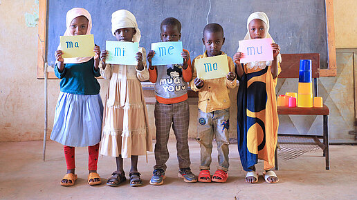 Fünf Grundschulkinder stehen nebeneinander vor einer Tafel und halten Schilder in der Hand.