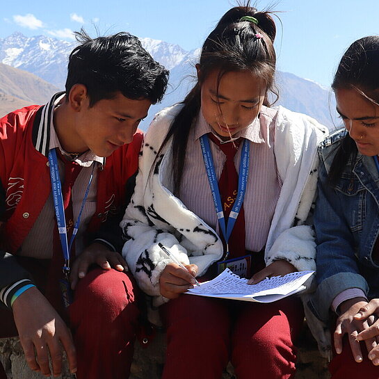 Drei Jugendliche sitzen auf einer Mauer. Das Mädchen in der Mitte schreibt in ein Heft. Im Hintergrund sieht man die Berge des Himalayas.