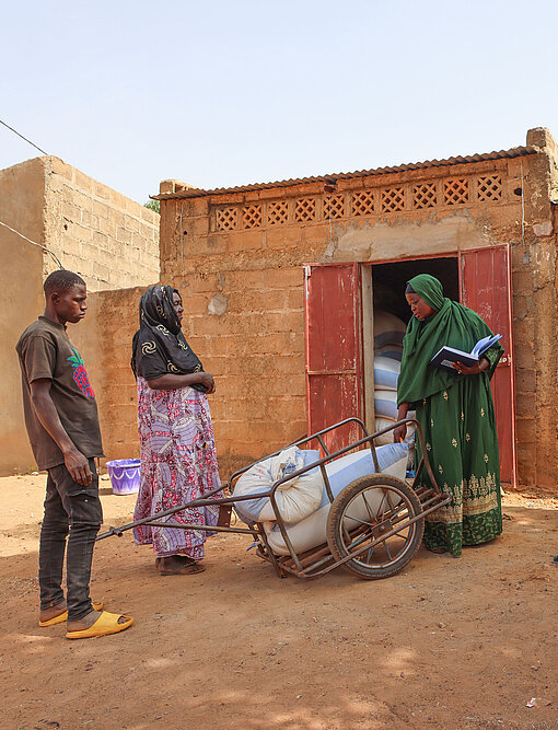 Frau liefert Mais an die Getreidebank, eine Frau mit Buch kontrolliert, drei Frauen sitzen daneben
