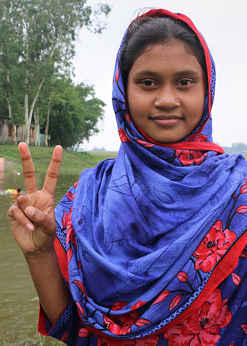 Ein Mädchen mit buntem Kleid und Kopftuch steht vor einem Teich und macht mit ihrer Hand das Peace-Zeichen.