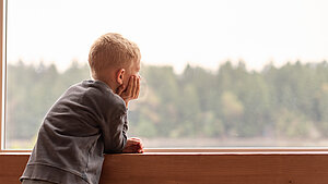  Junge sitzt auf einer Fensterbank und schaut nachdenklich in die Ferne auf einen Wald