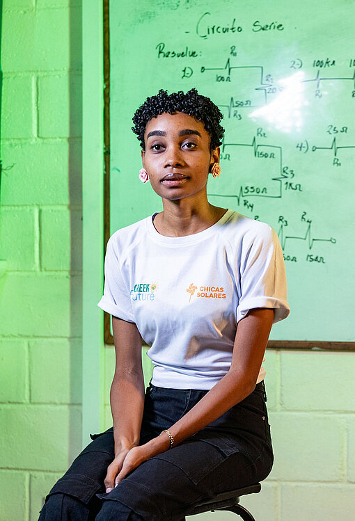 Porträt einer weiteren Projektteilnehmenden im Schulungsraum; sie sitzt vor einer beschrifteten Tafel mit Schaltplänen und trägt ein T‑Shirt des Ausbildungsprogramms.