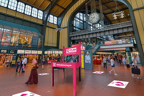 In einer Halle im Hamburger Hauptbahnhof steht ein überdimensional großer, pinker Stuhl mit der weißen Aufschrift "Plan International" und "weltmädchentag.de". Auf dem Boden daneben sind pinke Fußabdrücke auf weißem Grund geklebt, im Hintergrund laufen einige Menschen flüchtig vorbei.