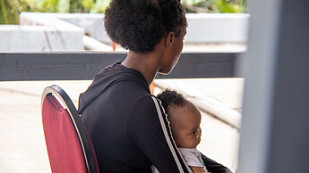 Eine Frau sitzt auf einem Stuhl und hat ein Baby auf dem Schoß. Sie sind von der Seite zu sehen. Die Frau schaut weg.