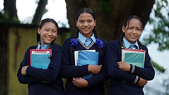 Drei nepalesische Mädchen in Schuluniform halten ihre Hefte in ihren Häden und lächeln