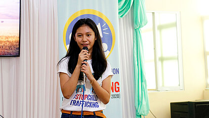 Eine junge Frau steht mit einem Mikrofron in einem Raum, im Hintergrund ist der Schriftzug "Stop Trafficking" zu erkennen.
