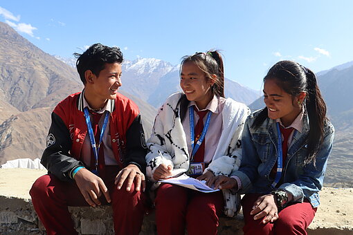 Drei Jugendliche sitzen auf einer Mauer und unterhalten sich. Im Hintergrund sieht man die Berge des Himalaya