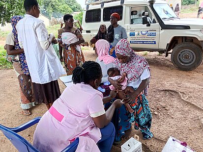 Mehrere Frauen und Kinder stehen im Freien neben einem weißen Geländewagen. Eine sitzende Person berührt das Bein eines Kleinkindes auf dem Schoß einer Frau. Weitere Personen stehen im Hintergrund, einige halten Kinder auf dem Arm.