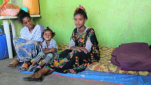 Sitzende Familie in einfachem Innenraum Drei Personen sitzen gemeinsam auf einer Matte in einem einfachen Raum und schauen in die Kamera.