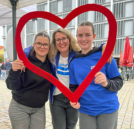 Zwei Mädchen und eine Frau blicken lächelnd durch die Silhouette eines großen roten Herzens in die Kamera