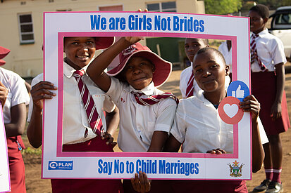 Drei Schülerinnen in Schuluniform halten gemeinsam einen Rahmen mit der Aufschrift „We are Girls Not Brides – Say No to Child Marriages“.