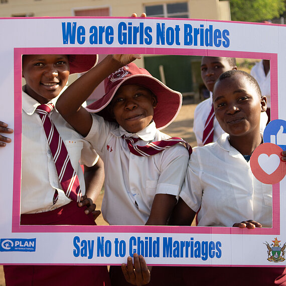 Drei Schülerinnen in Schuluniform halten gemeinsam einen Rahmen mit der Aufschrift „We are Girls Not Brides – Say No to Child Marriages“.