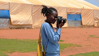 Clementine steht auf einem Platz und schaut durch den Sucher ihrer Kamera. Im Hintergrund ist ein großes Zelt.