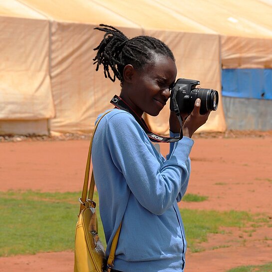 Clementine steht auf einem Platz und schaut durch den Sucher ihrer Kamera. Im Hintergrund ist ein großes Zelt.