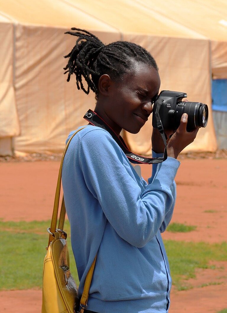 Clementine steht auf einem Platz und schaut durch den Sucher ihrer Kamera. Im Hintergrund ist ein großes Zelt.