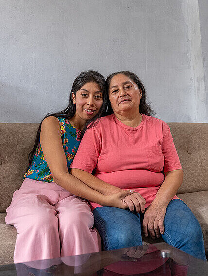 Mutter und Tochter sitzen gemeinsam auf einem Sofa und halten sich glücklich in den Armen; beide schauen gemeinsam in die Kamera.