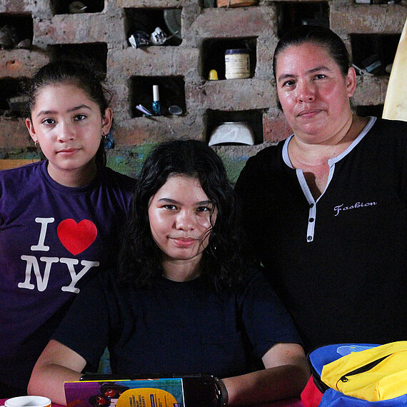 Drei Personen stehen zusammen in einem einfachen Raum mit Tisch und Surfbrett im Hintergrund – Symbolbild für Familien und Jugendliche in einem Projekt für Schutz und Bildung in El Salvador.