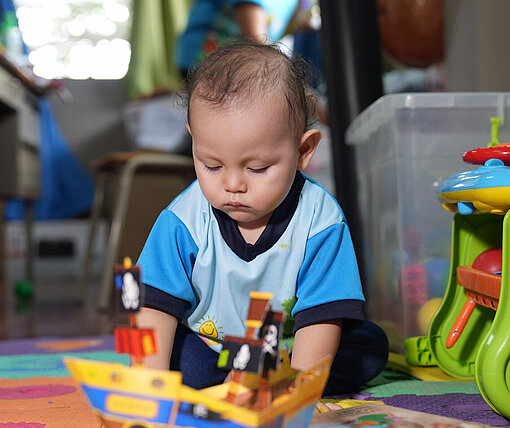 Von dem salvadorianischen Erziehungsansatz „Crecer juntos – Gemeinsam wachsen“ profitiert auch Derek Ein Kleinkind sitzt auf dem Boden zwischen bunten Spielsachen