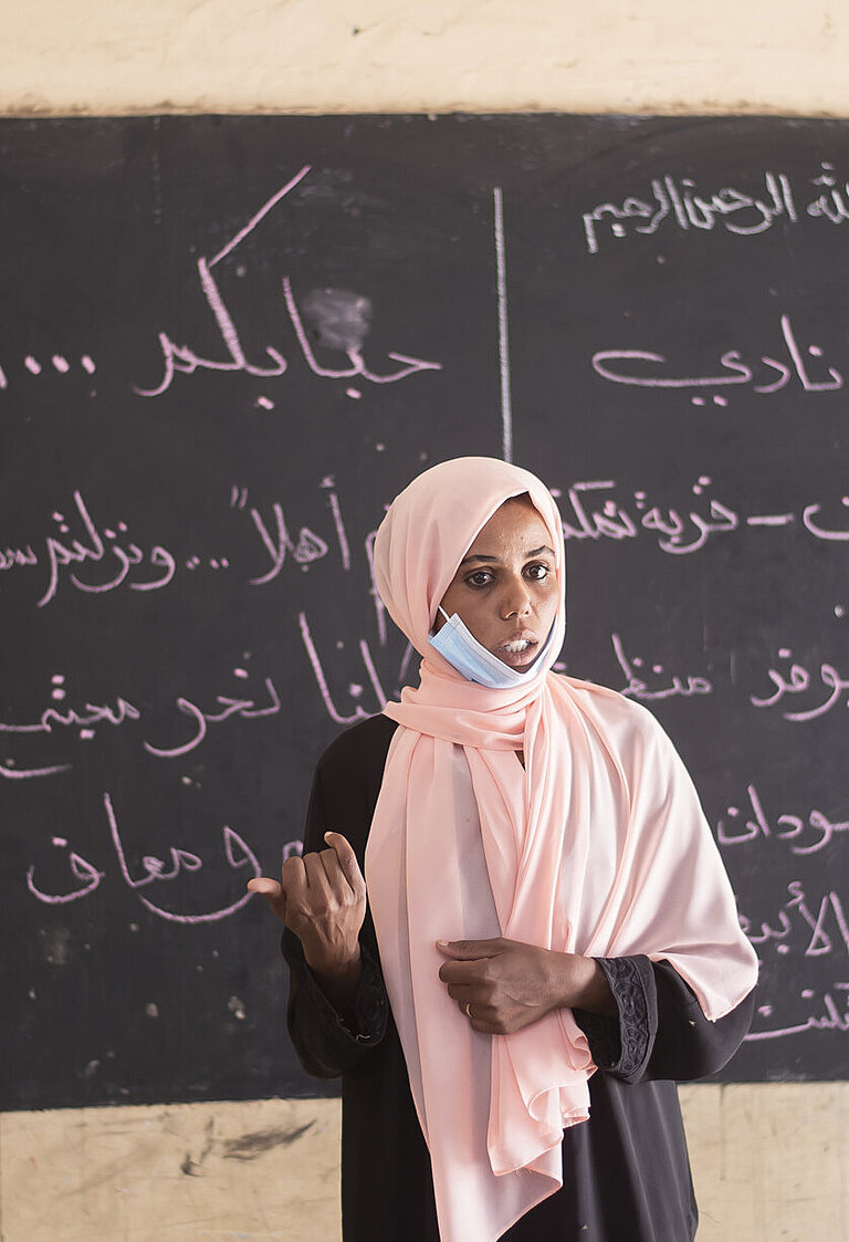Eine junge Frau mit Kopftuch steht vor einer beschriebenen Tafel und spricht.