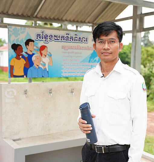 Ein Arzt steht vor Wasserspendern und hält eine Wasserflasche in der Hand, die er gerade aufgefüllt hat.