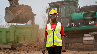 Eine Frau steht in Warnweste und mit Schutzhelm auf einer Baustelle, hinter ihr ist ein Bagger in Aktion