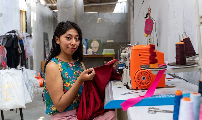 Eine junge Frau sitzt an einer Overlock-Nähmaschine und näht ein Kleidungsstück in ihrer Werkstatt.
