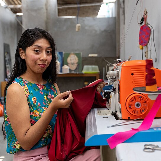 Eine junge Frau sitzt an einer Overlock-Nähmaschine und näht ein Kleidungsstück in ihrer Werkstatt.