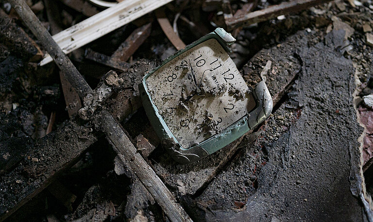 Eine kaputte Wanduhr liegt auf dem Boden, halb von Schutt und Asche verdeckt.