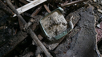 Eine kaputte Wanduhr liegt auf dem Boden, halb von Schutt und Asche verdeckt.