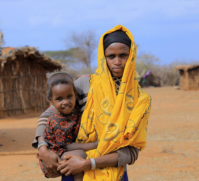 Eine Frau steht in einer trockenen Landschaft, sie hält ein Kind im Arm und schaut ernst in die Kamera