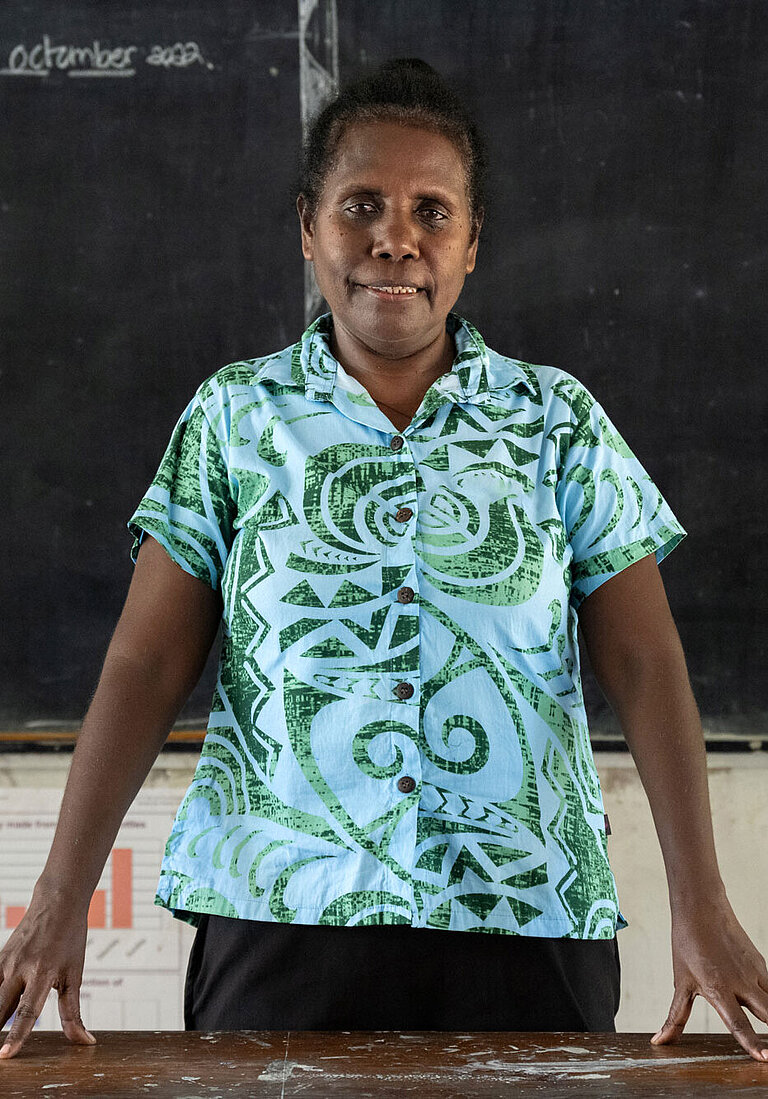 Eine Frau steht vor einer Tafel, hinter ihrem Pult in einem Klassenzimmer.