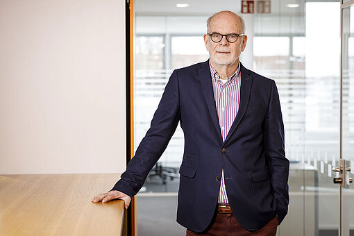 Dr. Axel Berger steht im Plan-Büro. Er trägt eine Anzugjacke und stützt sich locker auf einem Tisch ab.