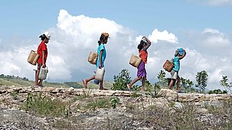 Eine Gruppe Kinder läuft über ein Feld und trägt Körbe mit Lasten auf ihren Köpfen
