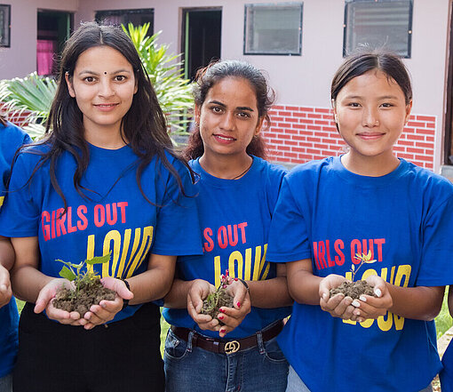Fünf Mädchen in blauen "Girls Out Loud"-T-Shirts halten kleine Pflanzen in den Händen