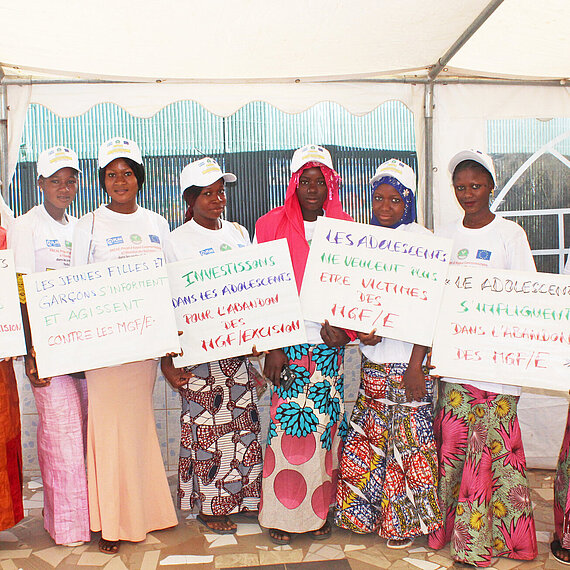 Junge Frauen und Mädchen in traditioneller Kleidung und weißen Plan T-Shirts stehen nebeneinander unter einem Zelt. Sie halten Schilder mit französischen Botschaften gegen weibliche Genitalverstümmelung.